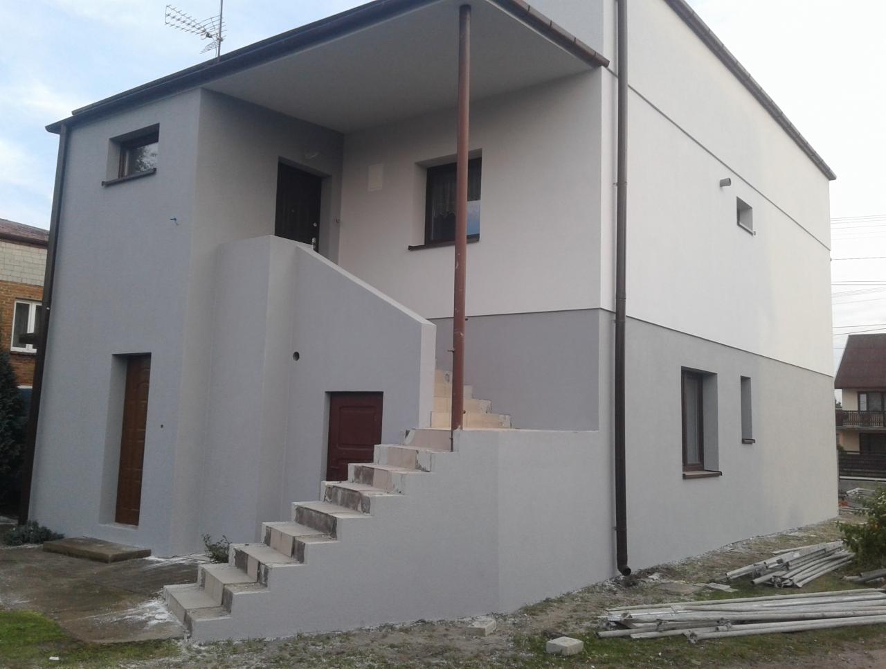 Dwukondygnacyjny dom po remoncie elewacji, z widocznymi schodami zewnętrznymi i świeżo położonym tynkiem w odcieniach szarości i bieli, rury spustowe w kolorze brązowym.