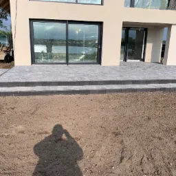 Nowoczesny taras z szarych płyt przed domem z dużymi przesuwnymi oknami, w tle widoczna koparka.