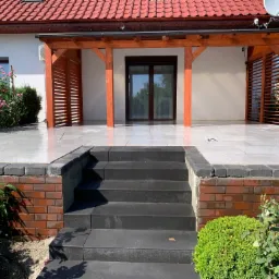 Nowoczesny taras z jasnych płyt, drewniana pergola i schody z ciemnej kostki granitowej, otoczone zielenią i różami.