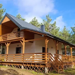 Nowoczesny dom z drewnianym tarasem i balkonem, otoczony zielenią drzew. Jasna elewacja kontrastuje z ciemnym dachem i drewnianymi elementami konstrukcyjnymi.