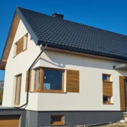 Nowoczesny dom jednorodzinny z białą elewacją, drewnianymi oknami i ciemną dachówką na tle błękitnego nieba i jesiennych drzew. Widoczny garaż z drewnianą bramą.