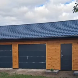 Drewniany garaż dwustanowiskowy z blachodachówką w Rybnie. Dwie bramy segmentowe i drzwi wejściowe w kolorze grafitowym. Elewacja z jasnego drewna.