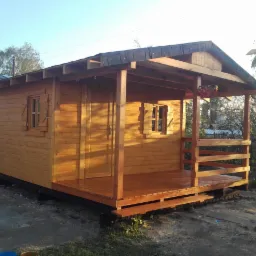 Drewniana konstrukcja zadaszonej werandy, świeżo pomalowana na brązowo, z widocznymi deskami i małymi oknami z okiennicami, otoczona zielenią i innymi budynkami.