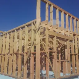 Wznoszenie drewnianej konstrukcji domu szkieletowego na fundamencie, widoczne elementy konstrukcyjne i stężenia tymczasowe, błękitne niebo w tle.