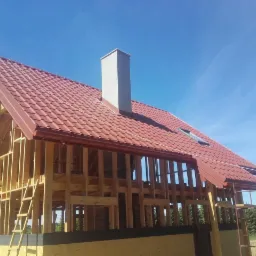Konstrukcja domu w trakcie budowy z widocznym drewnianym szkieletem, pokrytym częściowo czerwoną dachówką i szarym kominem na tle błękitnego nieba.