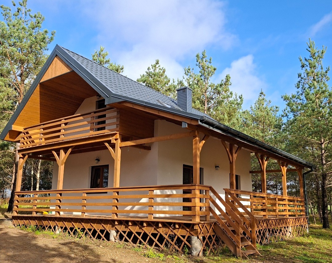 Nowoczesny dom z drewnianym tarasem i balkonem, otoczony zielenią drzew. Jasna elewacja kontrastuje z ciemnym dachem i drewnianymi elementami konstrukcyjnymi.