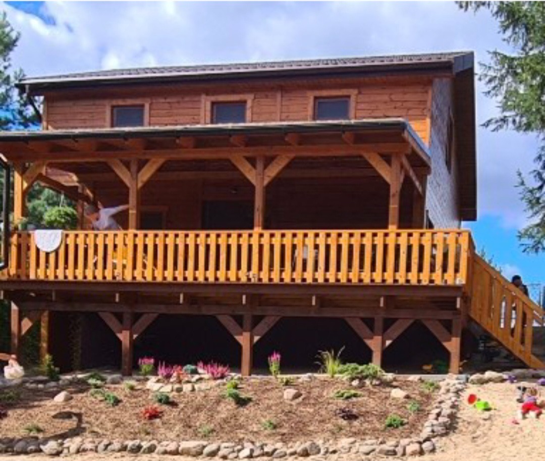 Drewniany domek letniskowy z tarasem i balustradą, otoczony rabatami kwiatowymi i zielenią. Widoczne elementy konstrukcyjne i schody. Jasne, naturalne oświetlenie.