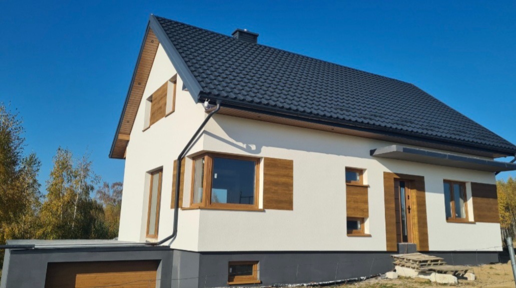 Nowoczesny dom jednorodzinny z białą elewacją, drewnianymi oknami i ciemną dachówką na tle błękitnego nieba i jesiennych drzew. Widoczny garaż z drewnianą bramą.