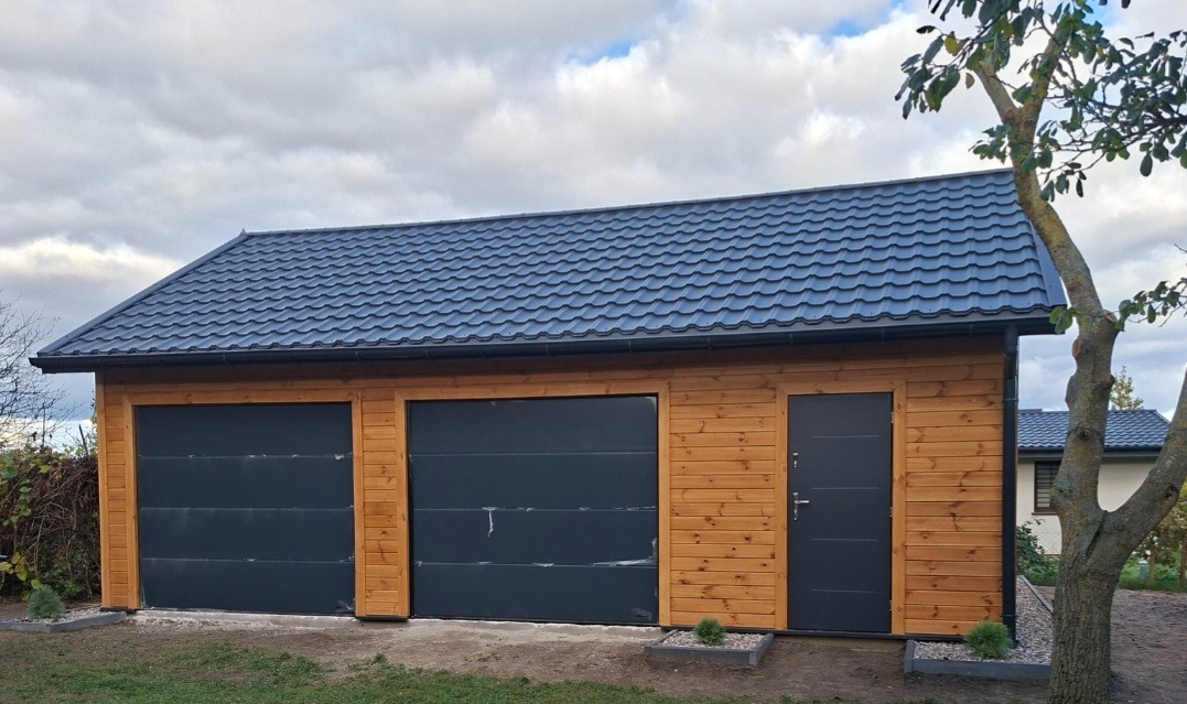 Drewniany garaż dwustanowiskowy z blachodachówką w Rybnie. Dwie bramy segmentowe i drzwi wejściowe w kolorze grafitowym. Elewacja z jasnego drewna.