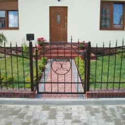 Ozdobne, czarne ogrodzenie z kutego żelaza z bramką wejściową, na fundamencie z cegły klinkierowej, prowadzące do domu z brązowymi oknami i drzwiami.