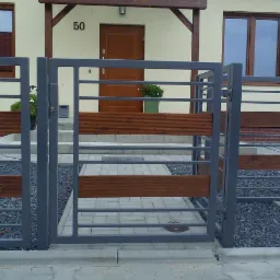 Szara metalowa furtka z poziomymi wstawkami z drewna, prowadząca do domu z numerem 50, widok z zewnątrz.