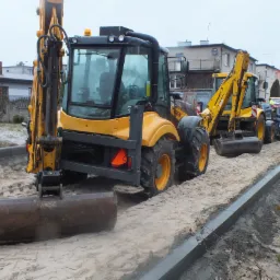 Dwa żółte traktory z łyżkami na piaszczystym podłożu obok ułożonych betonowych obrzeży, w tle zabudowania mieszkalne.