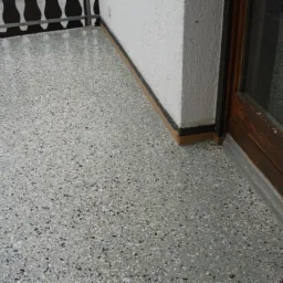Szara, gładka powierzchnia podłogi z drobnymi, ciemnymi plamkami, widoczna przy drzwiach balkonowych i białej ścianie.