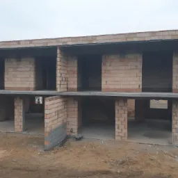 Dwukondygnacyjny budynek w stanie surowym otwartym, z widocznymi ścianami z cegły ceramicznej i betonowym stropem, na placu budowy z widoczną ziemią.