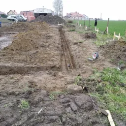 Wykonany wykop pod fundament z ułożonym zbrojeniem, widoczne sznurki wyznaczające granice, ziemia wokół wykopu, w tle budynki mieszkalne i zaparkowany bus.