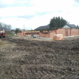 Wznoszenie ścian parterowego domu z czerwonej cegły, widoczny traktor i paleta z cegłami na ujeżdżonej działce budowlanej.