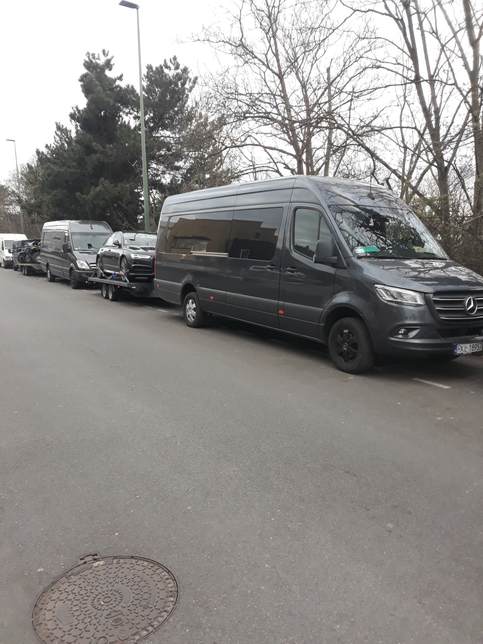 Szara laweta z czarnym Audi A8 na przyczepie, za nią kilka innych lawet z pojazdami, ustawione na ulicy, drzewa w tle.