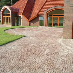 Ułożona kostka brukowa z różowego granitu tworząca podjazd do domu z czerwoną dachówką, widoczne zielone trawniki.