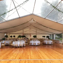 Uroczysty namiot z drewnianą podłogą, okrągłymi stołami z białymi obrusami i złotymi krzesłami, dekorowany zielonymi girlandami i żyrandolami, przygotowany na przyjęcie.