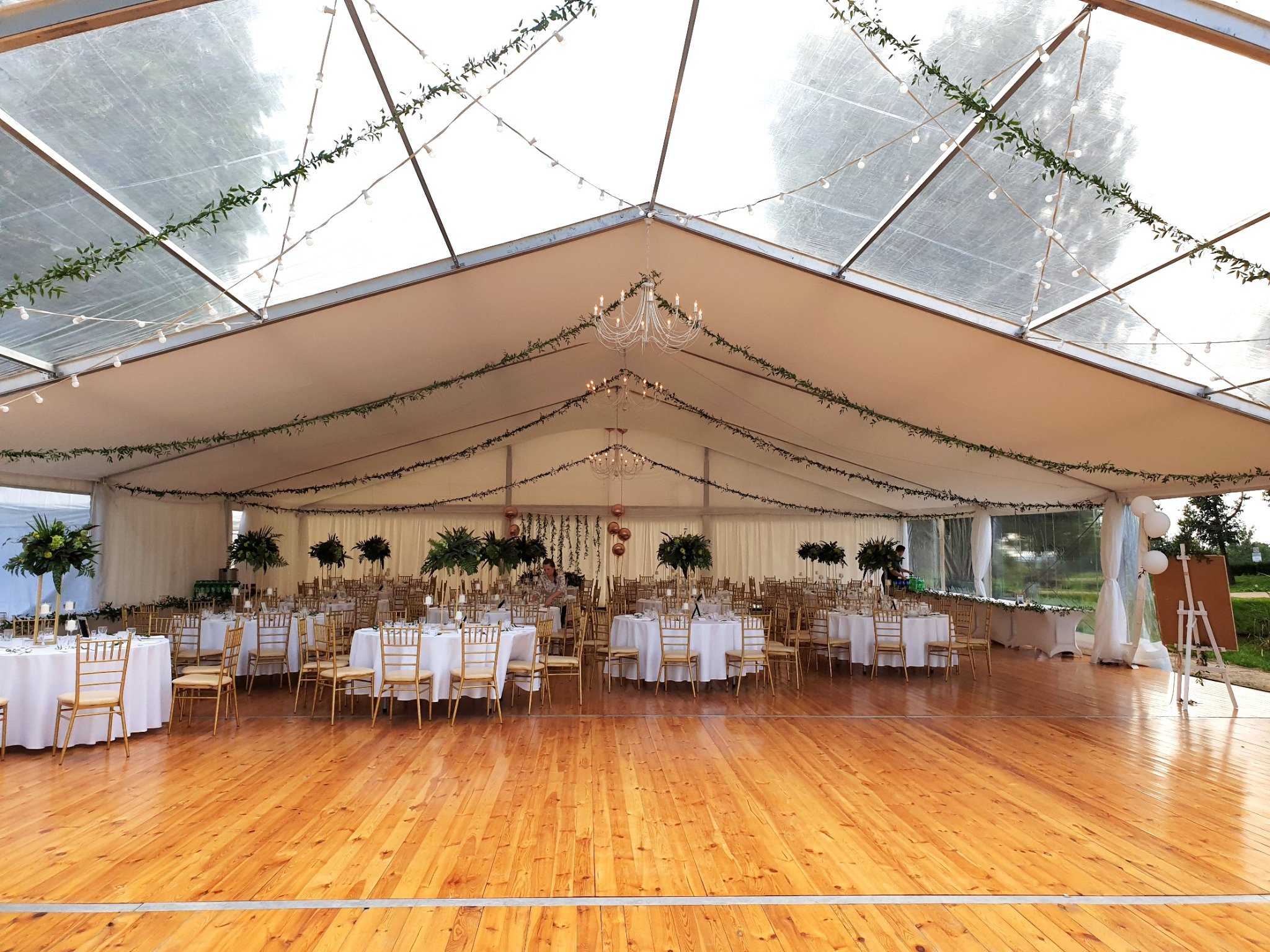Uroczysty namiot z drewnianą podłogą, okrągłymi stołami z białymi obrusami i złotymi krzesłami, dekorowany zielonymi girlandami i żyrandolami, przygotowany na przyjęcie.