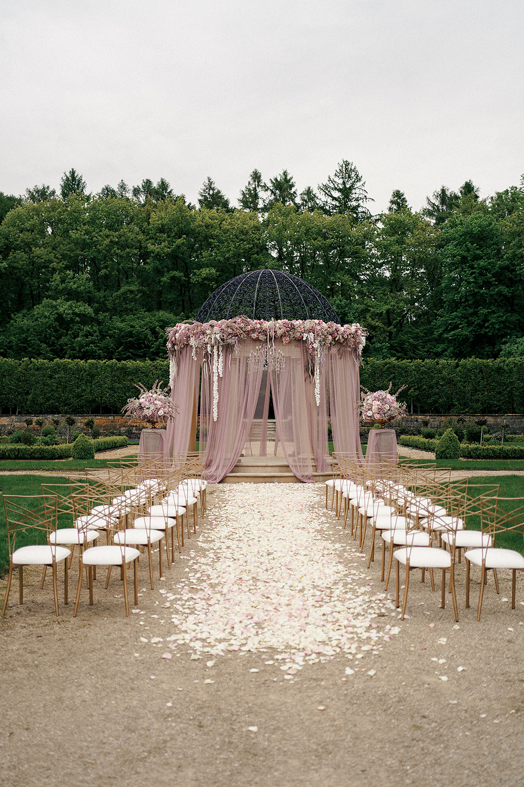 Uroczysta aranżacja ślubna na zewnątrz: metalowa altana z kwiatami i zwiewnymi zasłonami, ścieżka z płatków róż, rzędy złotych krzeseł z białymi poduszkami, zielone drzewa w tle.