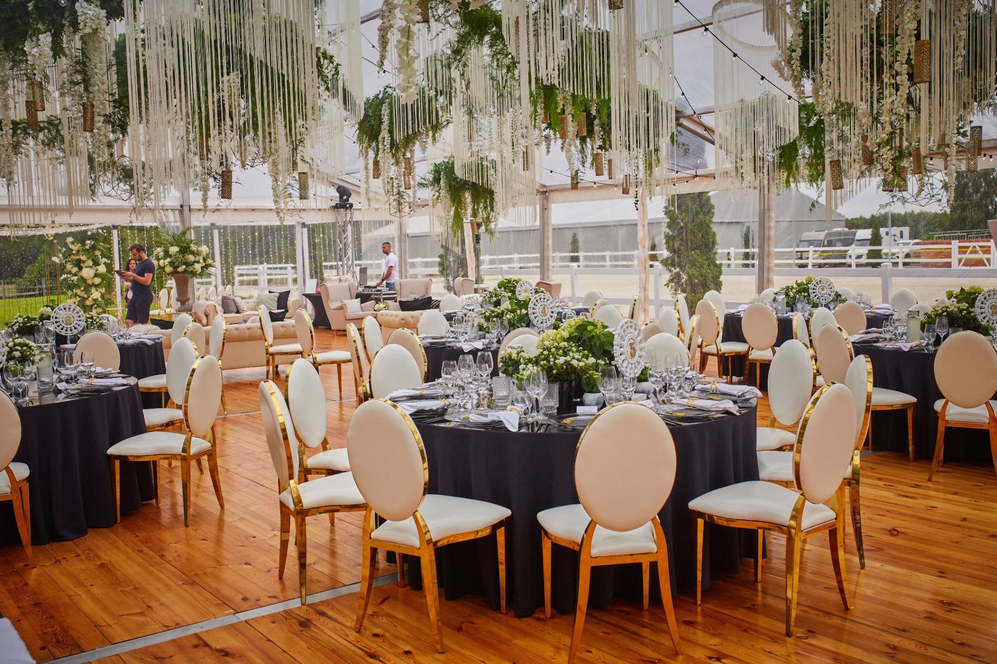 Uroczyste przyjęcie w namiocie: okrągłe stoły z czarnymi obrusami, biało-złote krzesła, kwiatowe dekoracje, zwisające ozdoby z roślin i białych wstążek.