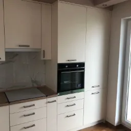 Jasna kuchnia z beżowymi frontami, zabudowana piekarnikiem, widoczne drewniane blaty i marmurowe wykończenie ściany nad blatem.