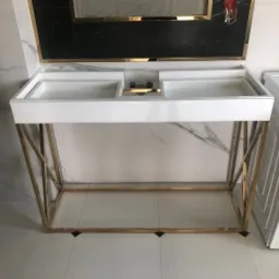 Biała, lakierowana umywalka z dwoma komorami na złotym, geometrycznym stelażu, na tle marmurowej ściany i czarno-białej podłogi, obok pralka.