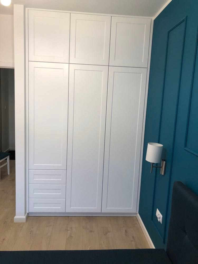 Biała szafa wnękowa z trzema szufladami na dole, dopasowana do niebieskiej ściany z dekoracyjnymi listwami i lampką ścienną.