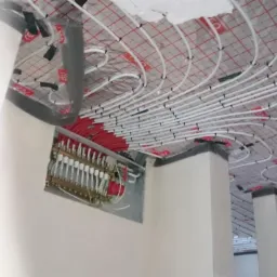 Rozłożona instalacja ogrzewania podłogowego na folii z siatką, widoczny rozdzielacz z rurami czerwonymi i białymi, wnętrze w trakcie prac wykończeniowych.