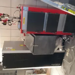 Czerwono-szary piec grzewczy z podłączonymi rurami, widoczna drabina oparta o piec, przewody elektryczne na podłodze, białe płytki na ścianie.