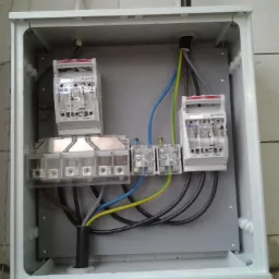 Rozdzielnica elektryczna w białej obudowie, widoczne bezpieczniki, listwy zaciskowe i kable w kolorowej izolacji, zamontowana na ścianie z bloczków betonowych.