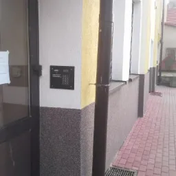 Zewnętrzny panel domofonu z klawiaturą numeryczną, zamontowany na elewacji budynku obok drzwi wejściowych, widoczna rynna i chodnik z kostki.