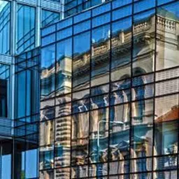 Szklana fasada nowoczesnego biurowca odbijająca klasyczną architekturę kamienicy w centrum miasta.