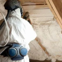 Pracownik w kombinezonie ochronnym aplikuje piankę izolacyjną na poddasze, widoczna warstwa pianki i drewniana konstrukcja dachu.