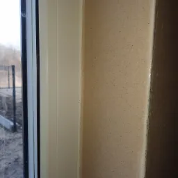 Fragment ściany z tynkiem maszynowym o fakturze baranka, przylegający do ramy okiennej z widokiem na zewnątrz.