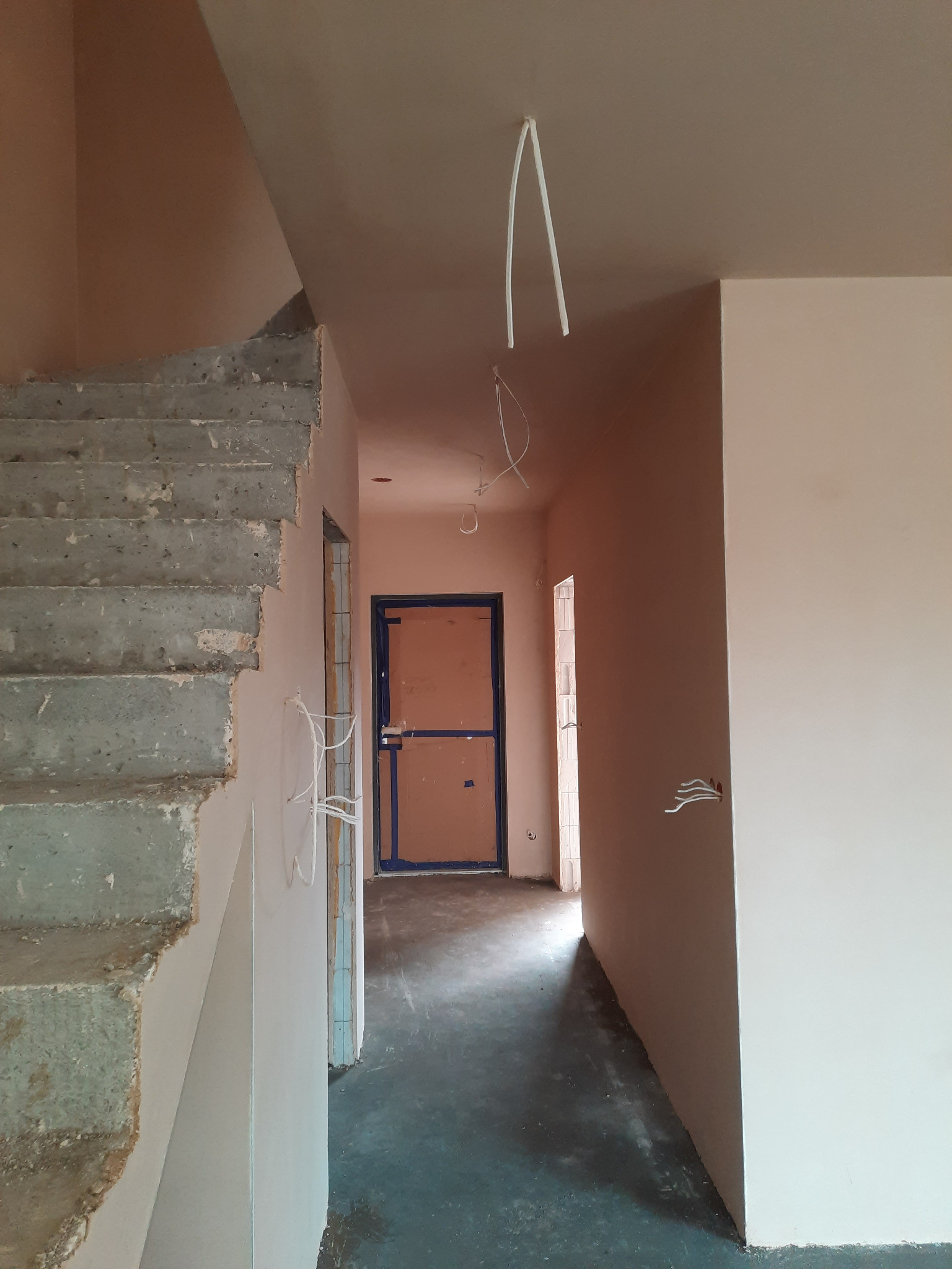 Wnętrze w trakcie prac wykończeniowych: surowe schody betonowe, świeżo otynkowane ściany w odcieniach beżu, wystające przewody elektryczne i niebieskie futryny drzwi w perspektywie korytarza.