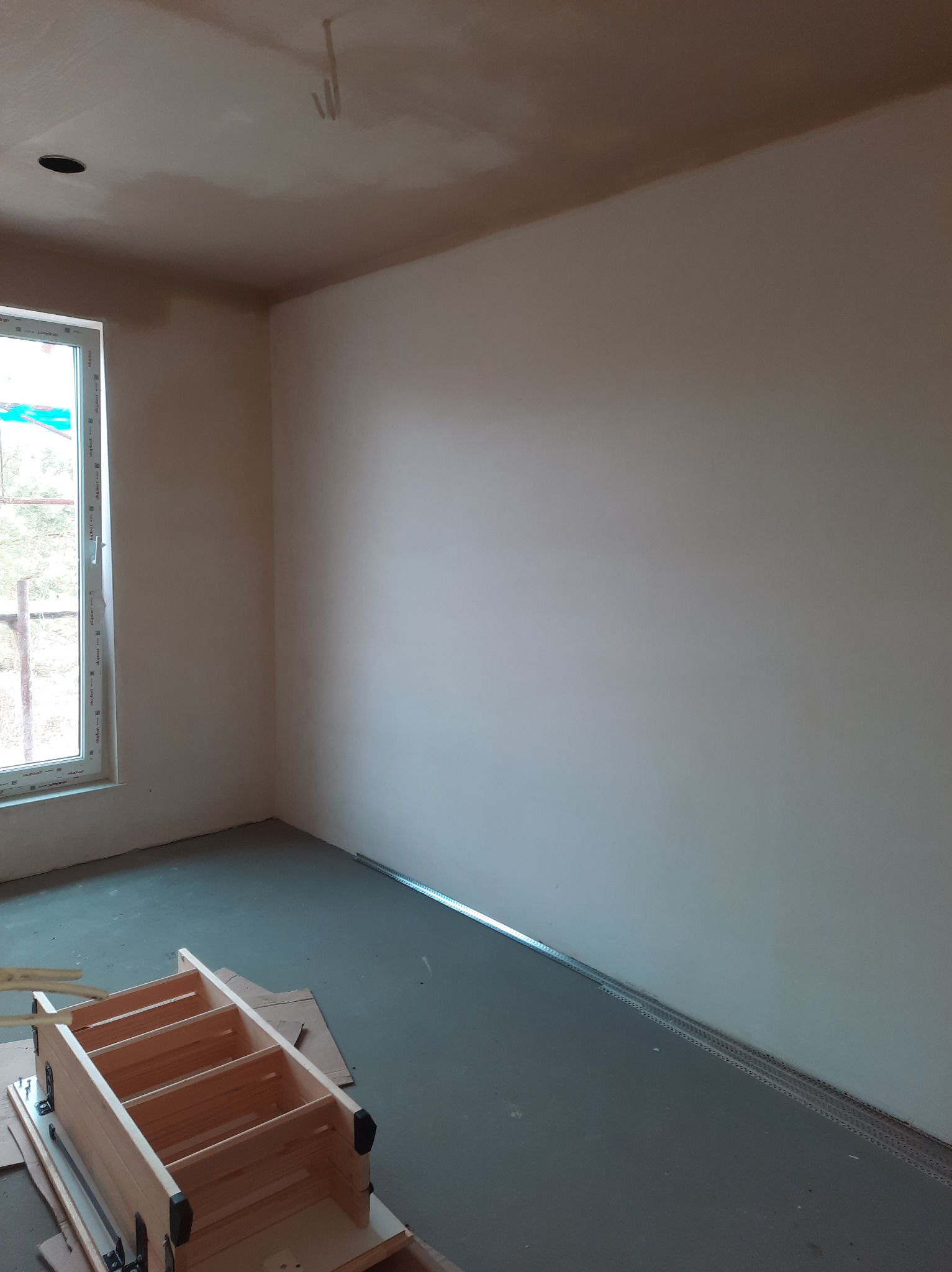 Wnętrze po nałożeniu jasnej, gładkiej warstwy gipsu na ścianach i suficie, widoczna listwa przypodłogowa i drewniana drabina składana w rogu pomieszczenia przy oknie.