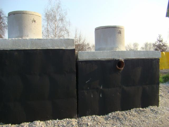 Dwa betonowe szamba z czarną izolacją, z widocznymi kominkami rewizyjnymi i rurą odpływową w jednym z nich, ustawione na żwirowym podłożu.