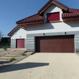 Dwukondygnacyjny dom z garażem i czerwoną dachówką. Biało-szara elewacja z poziomymi pasami. Podjazd wyłożony kostką, widoczne schody i rabaty.