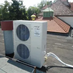 Biała jednostka zewnętrzna klimatyzacji MDV zamontowana na płaskim dachu, z widocznymi przewodami chłodniczymi i elementami instalacji, na tle budynku z czerwoną dachówką i zieloną wieżyczką.