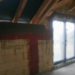 Wnętrze poddasza w trakcie prac murarskich: ściana z betonu komórkowego z widocznymi śladami zaprawy, częściowo zabezpieczone okno folią, drewniana konstrukcja dachu.