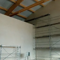 Wnętrze budynku w trakcie prac wykończeniowych, ściany pokryte świeżym tynkiem, widoczne rusztowania i drewniana konstrukcja dachu z blachą trapezową.