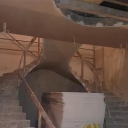 Wnętrze w trakcie prac wykończeniowych: surowe schody, rusztowania, stos płyt gipsowo-kartonowych i fragment nietypowego sufitu z otworem.