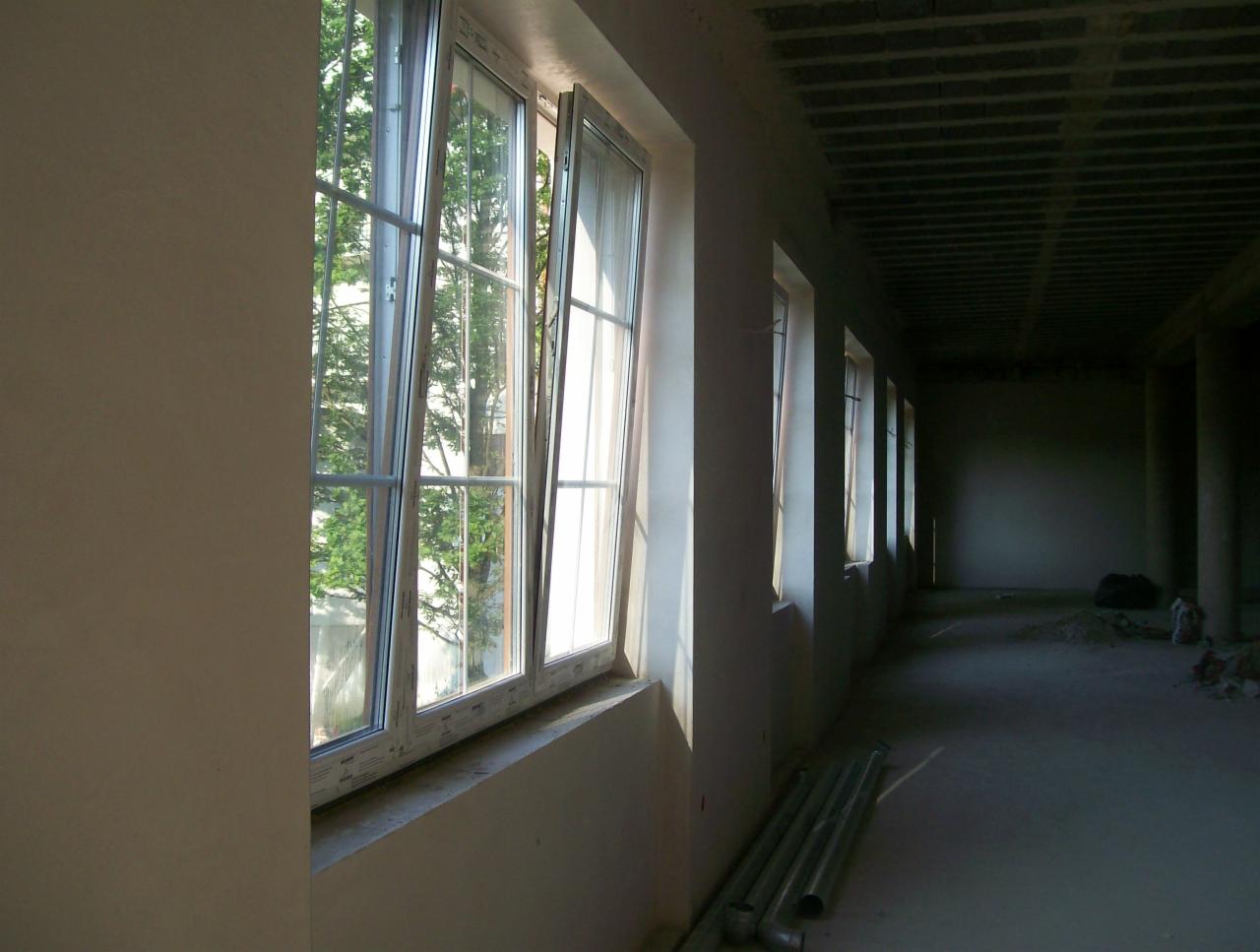 Wnętrze budynku w trakcie tynkowania: widok na okna z białymi ramami, częściowo otwarte, z widokiem na zieleń na zewnątrz; ściany w trakcie prac tynkarskich, surowy betonowy sufit, podłoga...