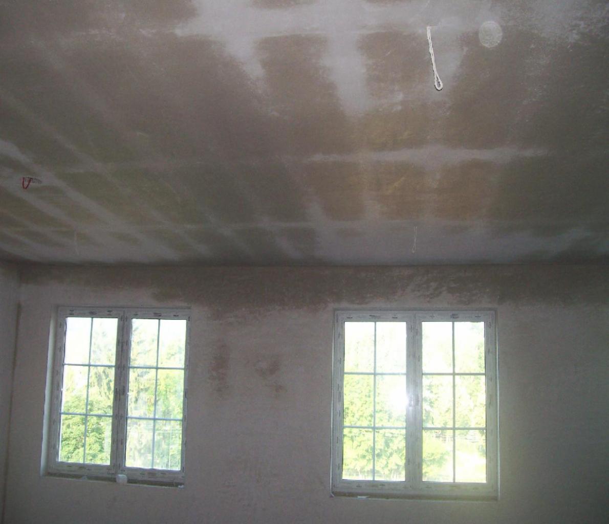 Wnętrze w Szczytnej w trakcie prac wykończeniowych: ściany i sufit po wstępnym nałożeniu gładzi, widoczne dwa okna z widokiem na zieleń.