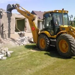 Żółta koparko-ładowarka JCB z łyżką kruszy resztki ściany budynku podczas prac rozbiórkowych, z gruzem leżącym na trawie. W tle widać fragment dachu innego budynku.