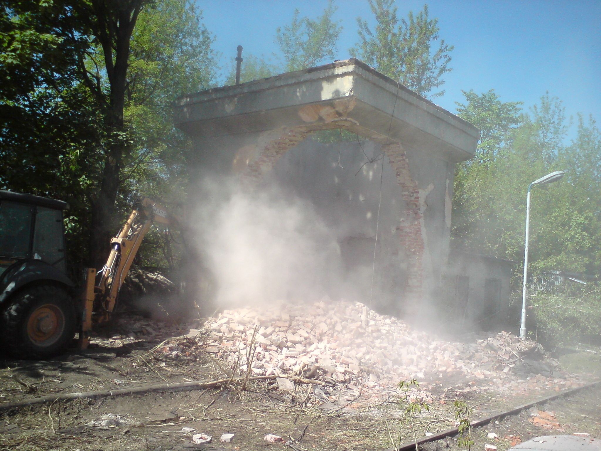 Częściowo zburzony budynek z cegły, otoczony pyłem, z widoczną koparką z boku i stertą gruzu na ziemi.