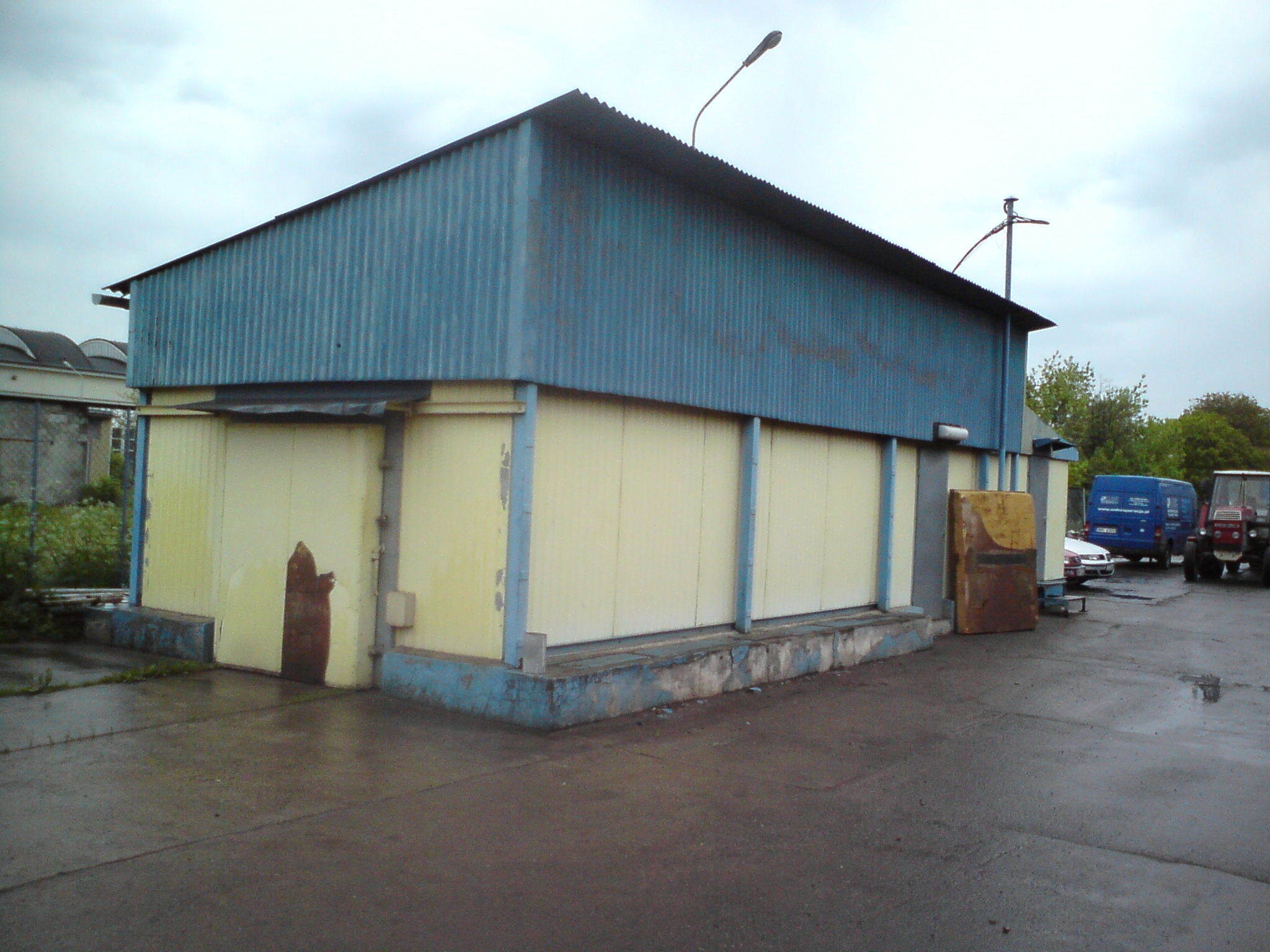 Niski, parterowy budynek magazynowy z blachy falistej w odcieniach niebieskiego i żółtego, z śladami rdzy na ścianie i podmokłym placem przed nim.