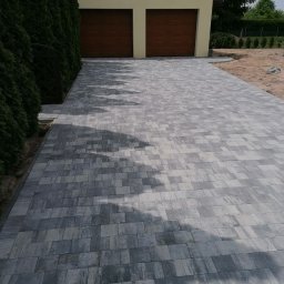 BRUK-MED Kamil Grądziel - Ułożona szara kostka brukowa prowadząca do garażu z drewnianymi drzwiami, widoczne cienie drzew na powierzchni, obok żywopłot z tui.